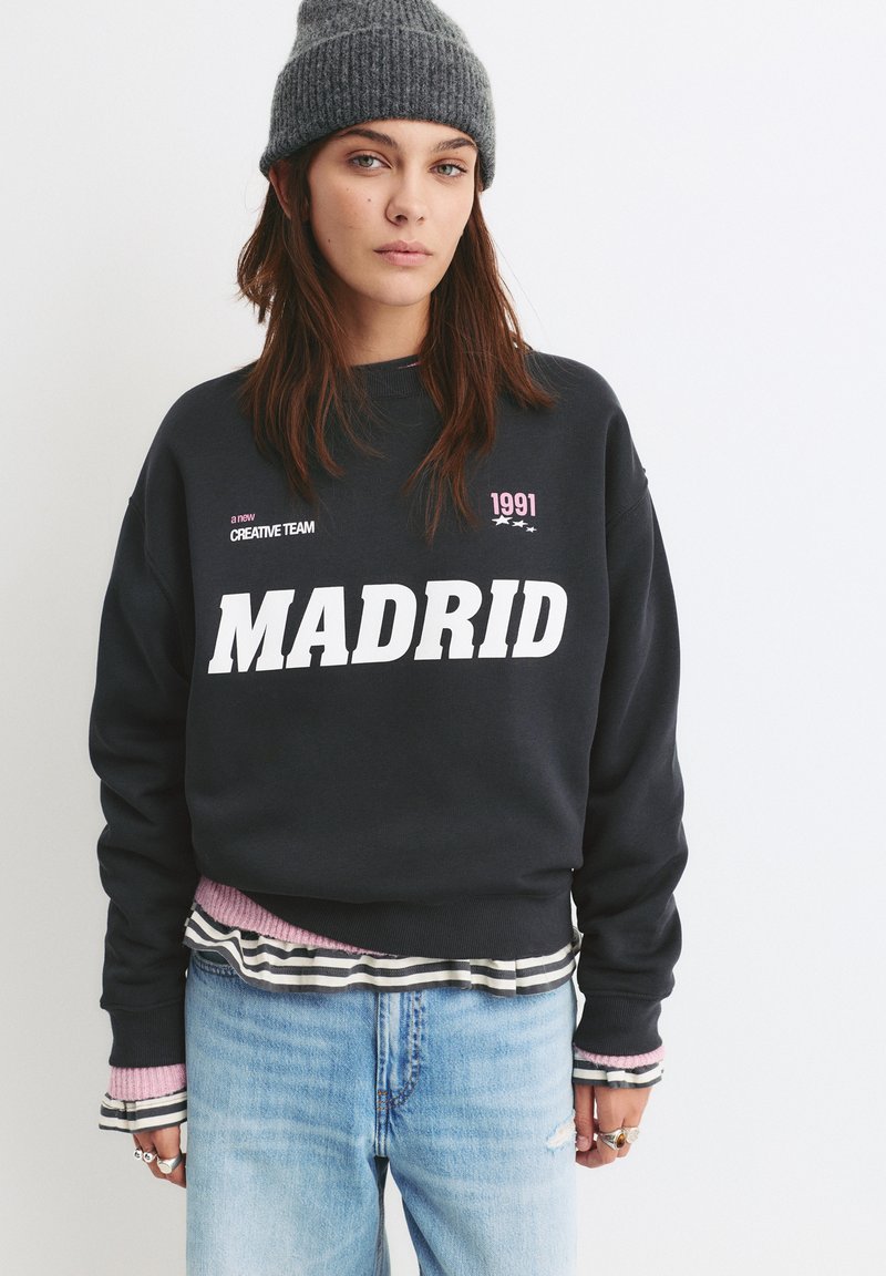 Námornícky sveter s veľkým bielym nápisom "MADRID", s ružovými akcentmi a pruhovanými vrstvenými manžetami, skombinovaný s svetlomodrými džínsami.