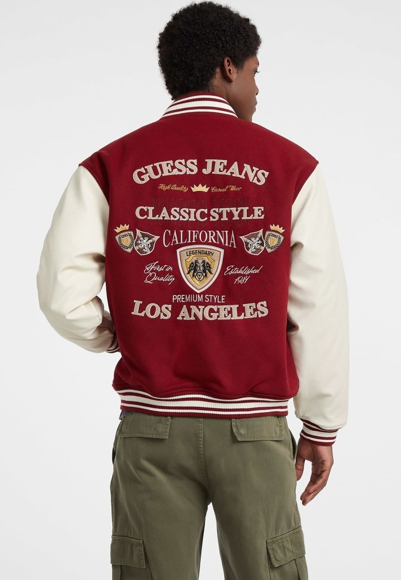 Punane ja kreemjas varsity jakk ribitud varrukatega, millel on broderitud graafika, sealhulgas "Guess Jeans" ja "Los Angeles" seljaosas.