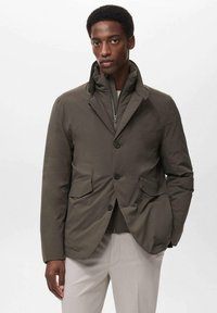 Man in een bruine knoopjack over een ritssluiting binnenlaag, in combinatie met lichtbeige broeken, staand tegen een effen achtergrond.