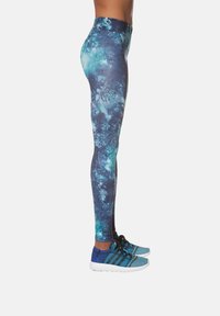 Legginsy z wysokim stanem w galaktyczny wzór w odcieniach niebieskiego i zielonego, wykonane z elastycznego materiału. Do tego niebieskie buty sportowe.