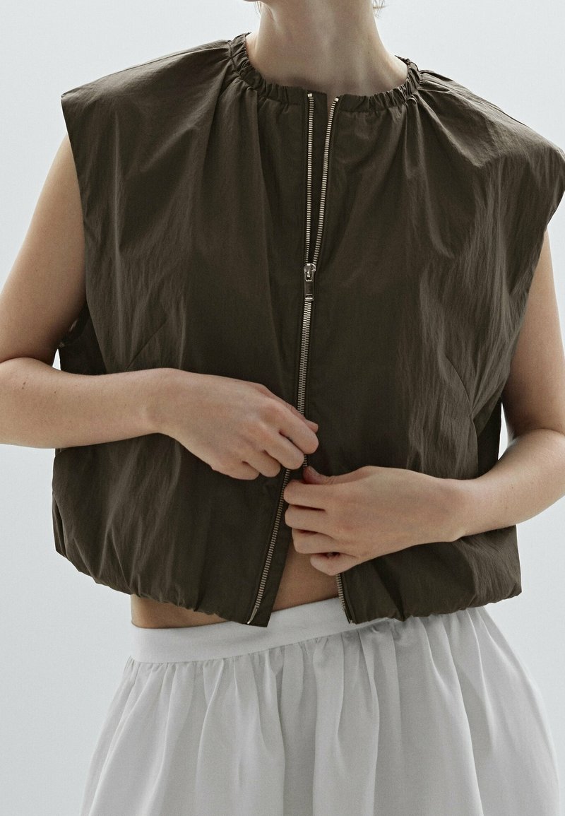 Personne portant une veste sombre sans manches avec fermeture éclair en train d'être zippée, associée à une jupe claire taille haute sur un fond neutre.