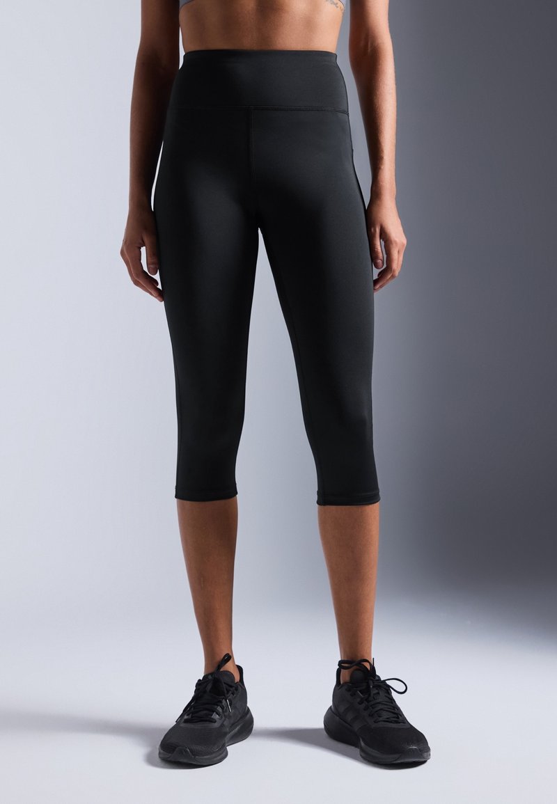 Personne portant des leggings de sport noirs taille haute et courts ainsi que des baskets noires, debout devant un fond neutre.