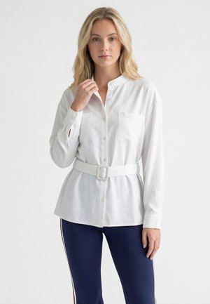 Donna con capelli biondi indossa una camicia bianca abbottonata con cintura e pantaloni blu scuro con righe laterali, in piedi davanti a uno sfondo chiaro e uniforme.