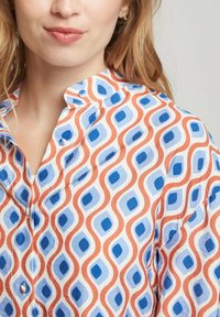 Chemise à boutons avec un motif de lignes ondulées orange et bleues et de carrés sur fond blanc ; tissu en coton, col et boutons visibles.
