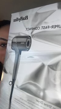 Person holder en Babyliss hårtørreræske, der viser en metalgrå hårtørrer mod en sølvfarvet krøllet baggrund.