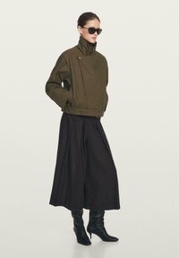 Giacca cropped verde oliva con colletto alto e tasche laterali, abbinata a pantaloni neri a gamba larga e stivali neri al ginocchio.
