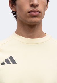 Νεαρός άνδρας με κρεμ Adidas crewneck μπλούζα, ορατός από τους ώμους μέχρι το πηγούνι, με ουδέτερη έκφραση, απλό ανοιχτόχρωμο φόντο.