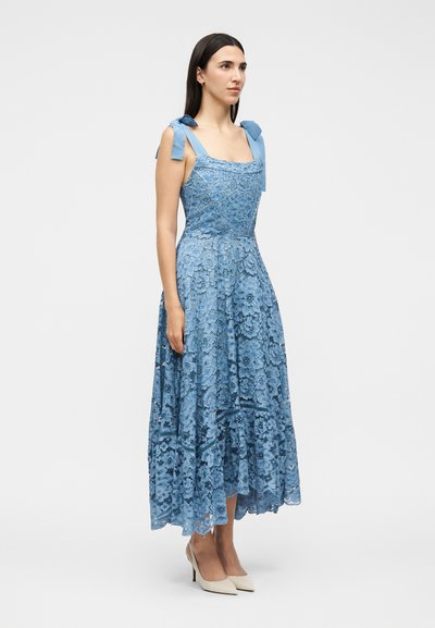 Γυναίκα με αμάνικο μπλε δαντελένιο φόρεμα μέχρι τους αστραγάλους με δεμένους ιμάντες στους ώμους και μπεζ ψηλοτάκουνα, στέκεται μπροστά σε λευκό φόντο.