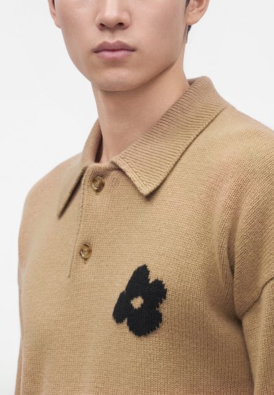 Homme portant un polo en maille beige avec deux boutons et un motif de fleur noire sur la poitrine, visible du cou jusqu’au milieu du torse.