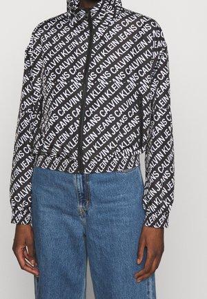 Musta vetoketjullinen takki, jossa valkoiset vinottain sijoitetut "Calvin Klein Jeans" -tekstit, yhdistetty sinisiin farkkuihin, kuvattu kaulasta lantioon.