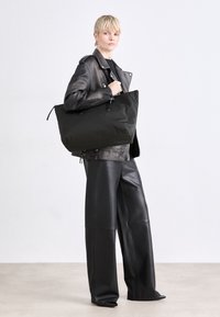 Černá oversized taška z texturované látky s dvojitými uchy a zipovým uzávěrem. Model nosí černou koženou bundu a široké kalhoty.