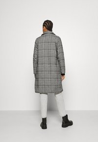 Persona con el pelo trenzado que lleva un abrigo largo de cuadros blanco y negro, pantalones gris claro y botas negras hasta el tobillo, de pie contra un fondo liso.