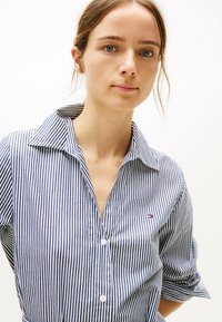 Camicia a righe blu e bianche con colletto ampio, dotata di un piccolo logo e maniche arrotolate. Realizzata in materiale di cotone leggero.