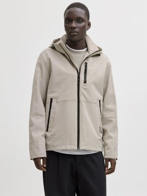 Ung mann iført en beige hettejakke med svarte glidelåsdetaljer og svarte bukser, står mot en ensfarget lys bakgrunn.