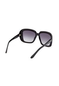 Lunettes de soleil carrées surdimensionnées noires avec verres dégradés foncés et logo "GUESS" sur la branche droite.