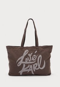 Ruskea kangaskassi pitkällä kahvalla, jossa on edessä hopeinen käsinkirjoitus "Love Karl". Pehmeä pinta ja tilava muotoilu tavaroiden kuljettamiseen.