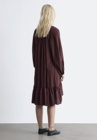 Bordeauxrotes Kleid mit langen Ärmeln, einem gerafften Saum und Rückendekolleté. Der Stoff sieht weich und fließend aus. Kombiniert mit schwarzen flachen Schuhen.