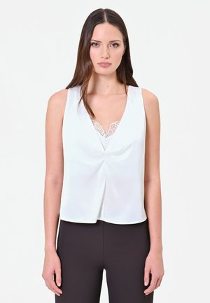 Femme aux longs cheveux bruns portant un haut blanc sans manches avec des détails en dentelle et un pantalon noir, debout devant un fond clair uni.