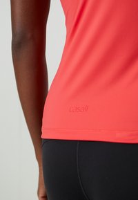 Coral athletic tank top with a smooth texture, featuring a subtle logo detail near the hem. Paired with black leggings.