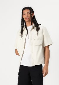 Cămașă crem cu mâneci scurte, cu nasturi în față, un buzunar la piept și branding subtil; purtată peste un tricou alb și pantaloni negri.