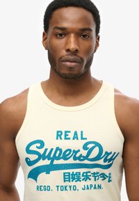 Jasno beżowy top na ramiączkach z wyraźnym niebieskim nadrukiem graficznym, na którym widnieje napis "REAL SuperDry" z dodatkowymi tekstami w kontrastowym stylu.