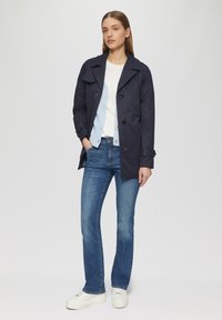 Marineblauwe trenchcoat met grote knopen, zijzakken en schouderepauletten, gecombineerd met lichte denim jeans en witte sneakers.