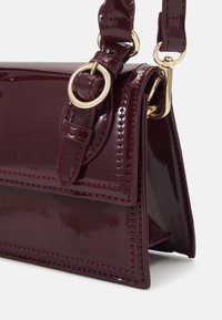 Burgundy patent leather bag with gold hardware, featuring a structured shape and visible stitching details. Adjustable strap included.
