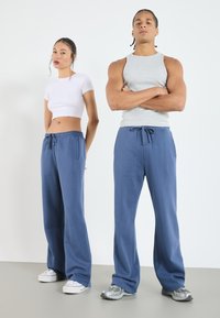 Blå vida träningsbyxor med elastisk midja och dragsko. Modellen bär en vit cropped topp; en annan modell har på sig en grå linne.