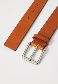 Ceinture en cuir tan présentant plusieurs trous pour réglage, une boucle en métal argenté poli, et une signalétique visible sur la boucle.