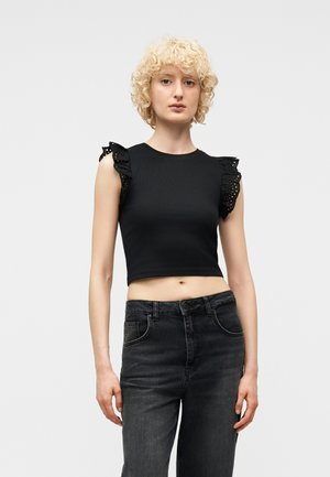 Ung kvinde med krøllet blondt hår iført en sort ærmeløs crop top med flæset øjeskylning og højtaljede mørke jeans.