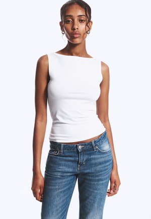 Ung kvinde med mørkt hår iført en hvid ærmeløs top og blå jeans, stående mod en ensfarvet lys baggrund.