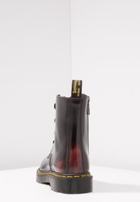 Černá kožená bojová bota s lesklým povrchem, žlutým prošíváním a smyčkou na horní části. Obsahuje zip a šněrování.