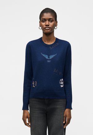 Kobieta ubrana w granatową bluzę z jasnoniebieską grafiką skrzydła, "1987", czaszkę i tekstowe detale, w połączeniu z ciemnoszarymi jeansami, stojąca.