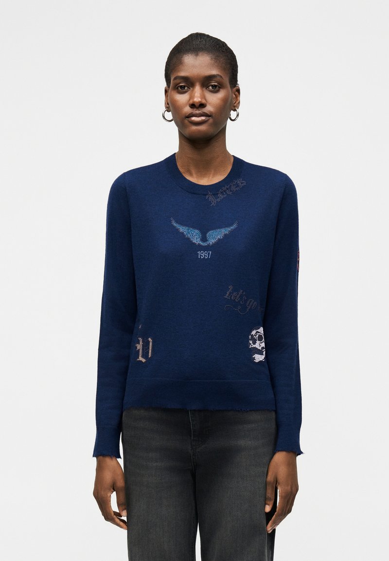 Γυναίκα που φοράει μπλε σκούρο φούτερ με σχέδιο φωτεινού μπλε φτερού, «1987», κρανίο και κείμενα, συνδυασμένο με σκούρα γκρι τζιν, όρθια.