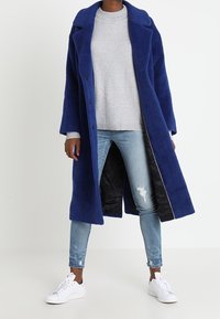 Blauer Wollmantel mit ausgeschlitztem Kragen, Knopfverschluss und seitlichen Taschen, getragen über einem grauen Pullover und hellblauen, verwaschenen Jeans. Weiße Sneakers.