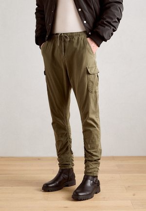 Olive green cargo pants with an elastic waistband, zipper, and pockets. Paired with brown ankle boots featuring a buckle detail.
