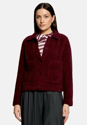 Vrouw draagt een bordeauxrode jas met knopen en grote voorzakken over een gestreepte trui en donkere wijde broek.