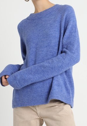 Kvinde iført en langærmet blå striktrøje og beige bukser, stående mod en ensartet hvid baggrund.