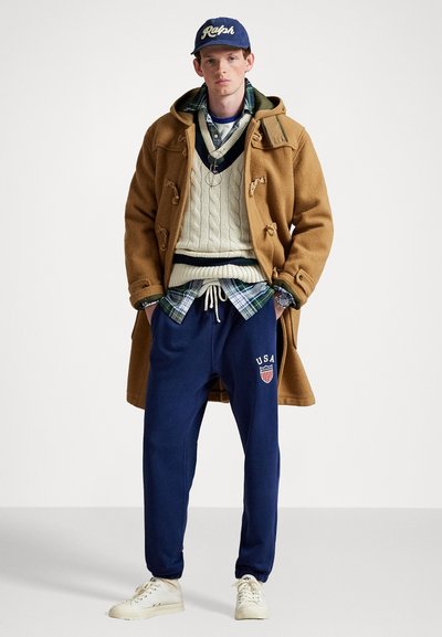 Palton din lână maro, pulover crem din tricot cu torsade, cămașă cu carouri albastre, pantaloni de trening bleumarin cu logo-ul "USA", adidași albi, șapcă albastră.