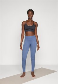 Musta urheiluliivi ohuilla olkaimilla ja logolla, yhdistettynä korkea فyxyn vaaleansiniset ribbileggingsit, seisoo joogamatolla.