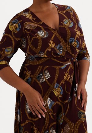 Frau trägt ein weinrotes Wickelkleid mit einem goldenen Ketten- und Vintage-Kofferetiketten-Muster, mit dreiviertellangen Ärmeln und gebundener Taille.