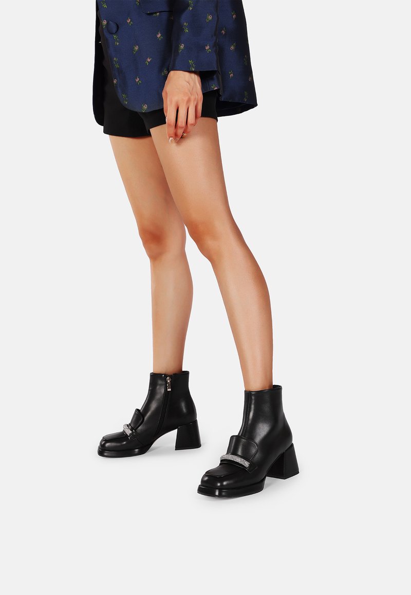 Stivali alla caviglia in pelle nera con tacco spesso, dotati di una fascia sulla parte anteriore e di una cerniera laterale. Texture liscia, design elegante.