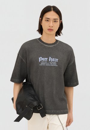 Junge Person mit glattem dunklem Haar trägt übergroßes graues "Pure Haze" T-Shirt und hält eine große schwarze Ledertasche vor einfachem Hintergrund.
