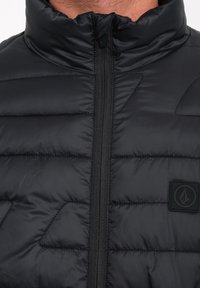 Zwarte gewatteerde vest met een hoge kraag, voorzien van een centrale ritssluiting en een gestructureerde logopatch aan de linkerzijde. Glad, glanzend materiaal.
