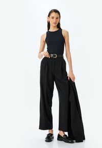 Top negro sin mangas, pantalones negros de cintura alta con pliegues, cinturón negro con hebilla dorada, zapatos negros, pose de pie, fondo minimalista.