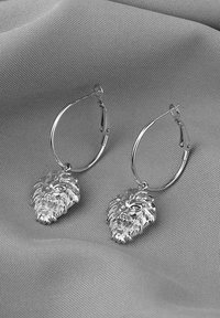 Silberne Creolenohrringe mit Löwenkopfmuster, detaillierte Mähnenstruktur und polierter Oberfläche, auf einem grauen Stoffhintergrund präsentiert.