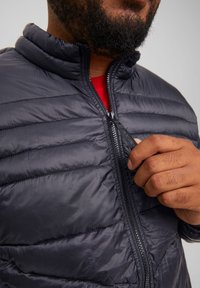 Chaqueta de plumas negra con cierre de cremallera, que presenta una superficie texturizada y acolchada, y un cuello alto. Mano ajusta la cremallera cerca del pecho.