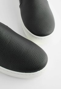 Chaussures en cuir noir à enfiler avec une finition texturée, semelles en caoutchouc blanches et coutures visibles le long de la semelle. Conception à bout arrondi.
