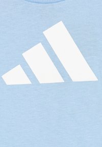 Camiseta de algodón azul claro con un gran diseño de logo de tres rayas blancas en la parte frontal. Textura suave, estilo de cuello redondo.