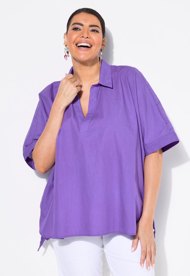 Camicia oversized viola con colletto e maniche corte, caratterizzata da un orlo spaccato e dettagli con bottoni sulle maniche. Tessuto liscio e leggero.
